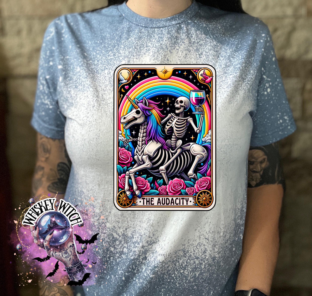 A Indigo blue   t-shirt with a bleached design featuring a tarot card with the image of  skeleton holding a glass of wine  riding a unicorn with a rainbow behind them with the words of “The Audacity” at the bottom