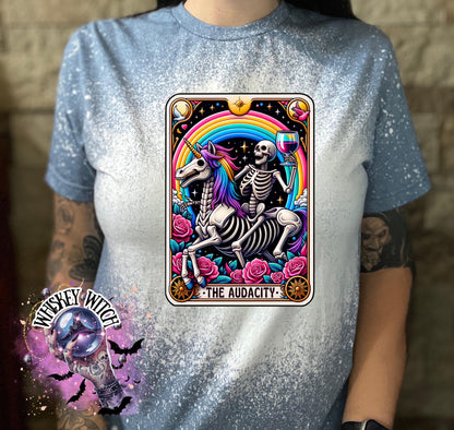 A Indigo blue   t-shirt with a bleached design featuring a tarot card with the image of  skeleton holding a glass of wine  riding a unicorn with a rainbow behind them with the words of “The Audacity” at the bottom