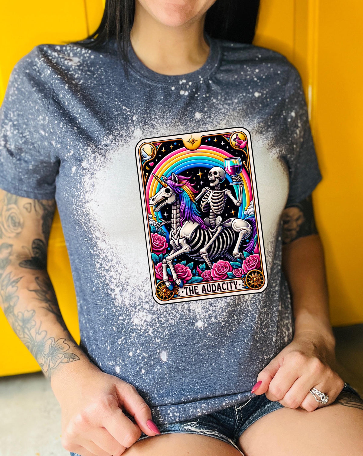 A navy blue   t-shirt with a bleached design featuring a tarot card with the image of  skeleton holding a glass of wine  riding a unicorn with a rainbow behind them with the words of “The Audacity” at the bottom