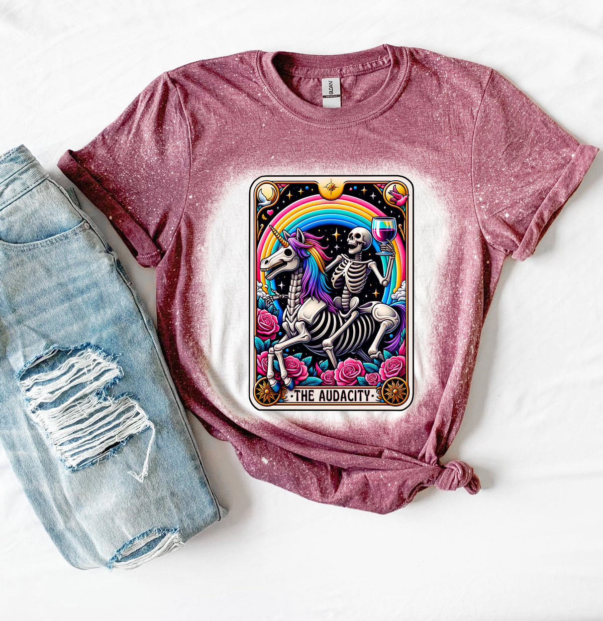 A maroon t-shirt with a bleached design featuring a tarot card with the image of  skeleton holding a glass of wine  riding a unicorn with a rainbow behind them with the words of “The Audacity” at the bottom