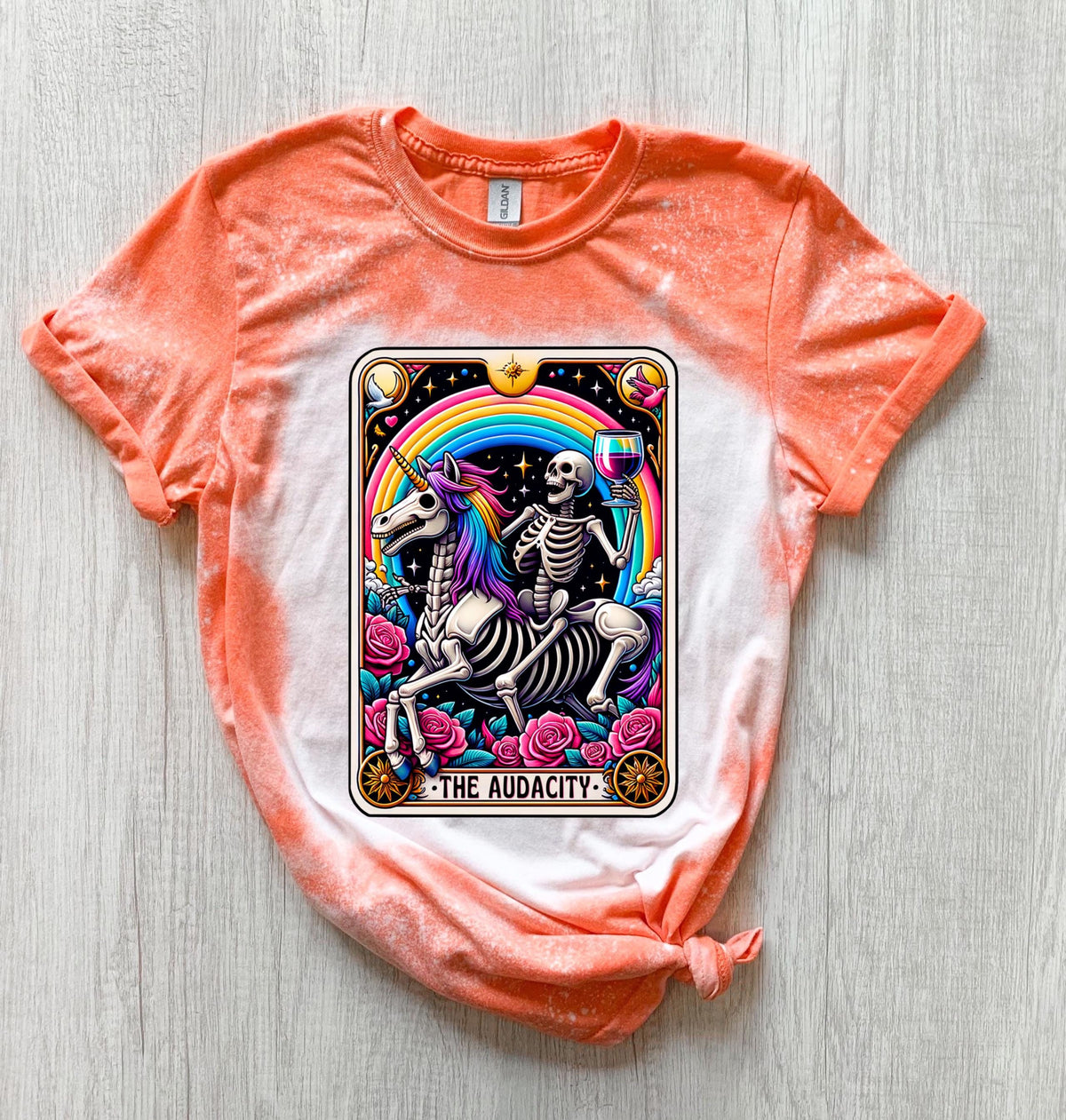 A Orange   t-shirt with a bleached design featuring a tarot card with the image of  skeleton holding a glass of wine  riding a unicorn with a rainbow behind them with the words of “The Audacity” at the bottom