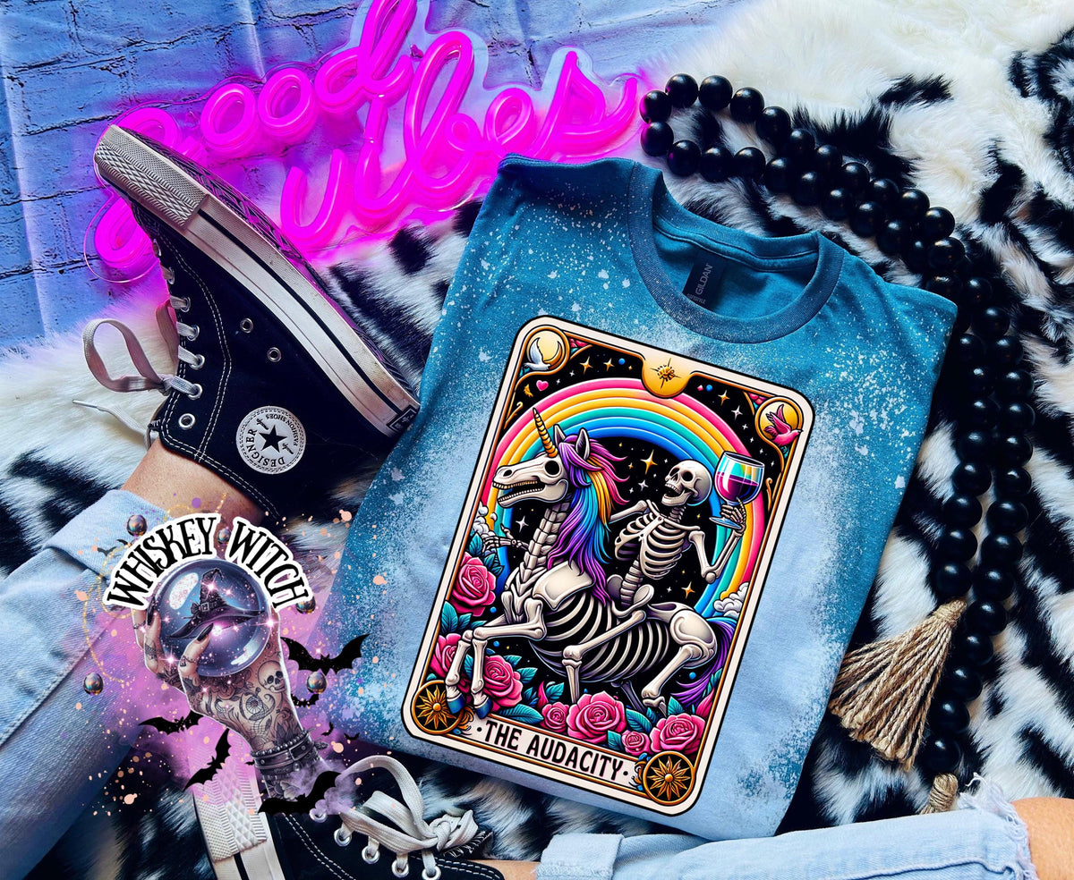 A Galapagos blue t-shirt with a bleached design featuring a tarot card with the image of  skeleton holding a glass of wine  riding a unicorn with a rainbow behind them with the words of “The Audacity” at the bottom