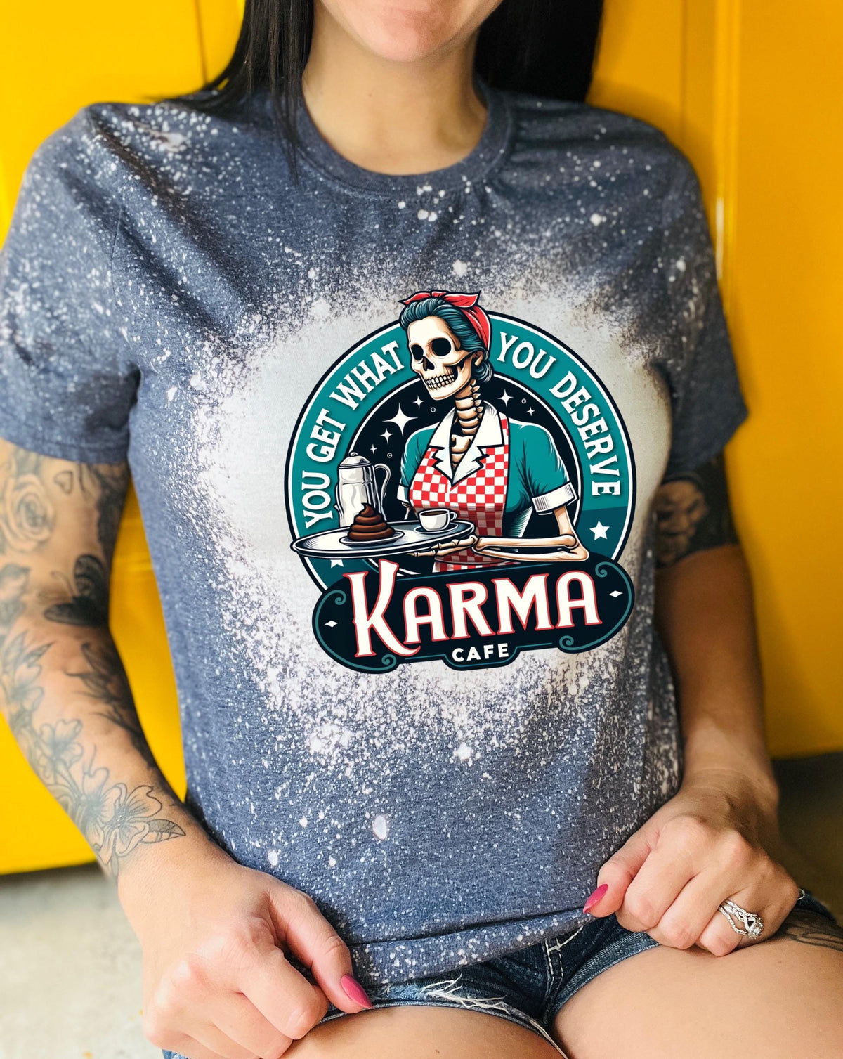 A navy blue  t-shirt with a bleached design featuring a skeleton wearing a red and white dress and holding a tray with a cup of coffee and a piece of cake. The text "You get what you deserve" is written around the skeleton. Below the skeleton is the text "Karma Cafe".