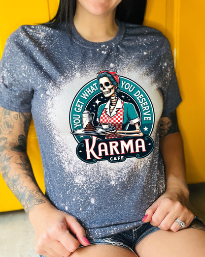 A navy blue  t-shirt with a bleached design featuring a skeleton wearing a red and white dress and holding a tray with a cup of coffee and a piece of cake. The text "You get what you deserve" is written around the skeleton. Below the skeleton is the text "Karma Cafe".