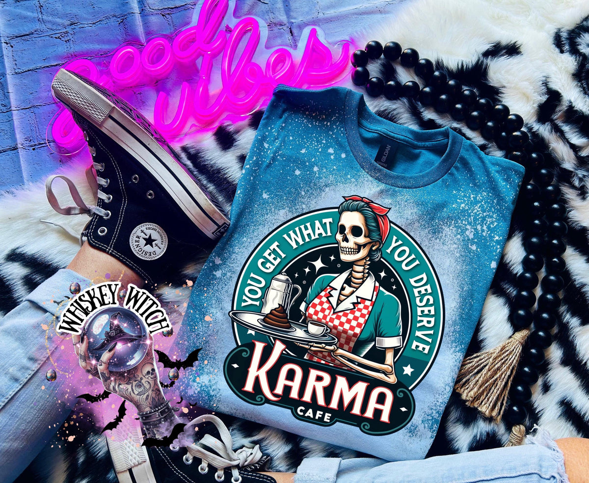 A Galapagos blue t-shirt with a bleached design featuring a skeleton wearing a red and white dress and holding a tray with a cup of coffee and a piece of cake. The text "You get what you deserve" is written around the skeleton. Below the skeleton is the text "Karma Cafe".