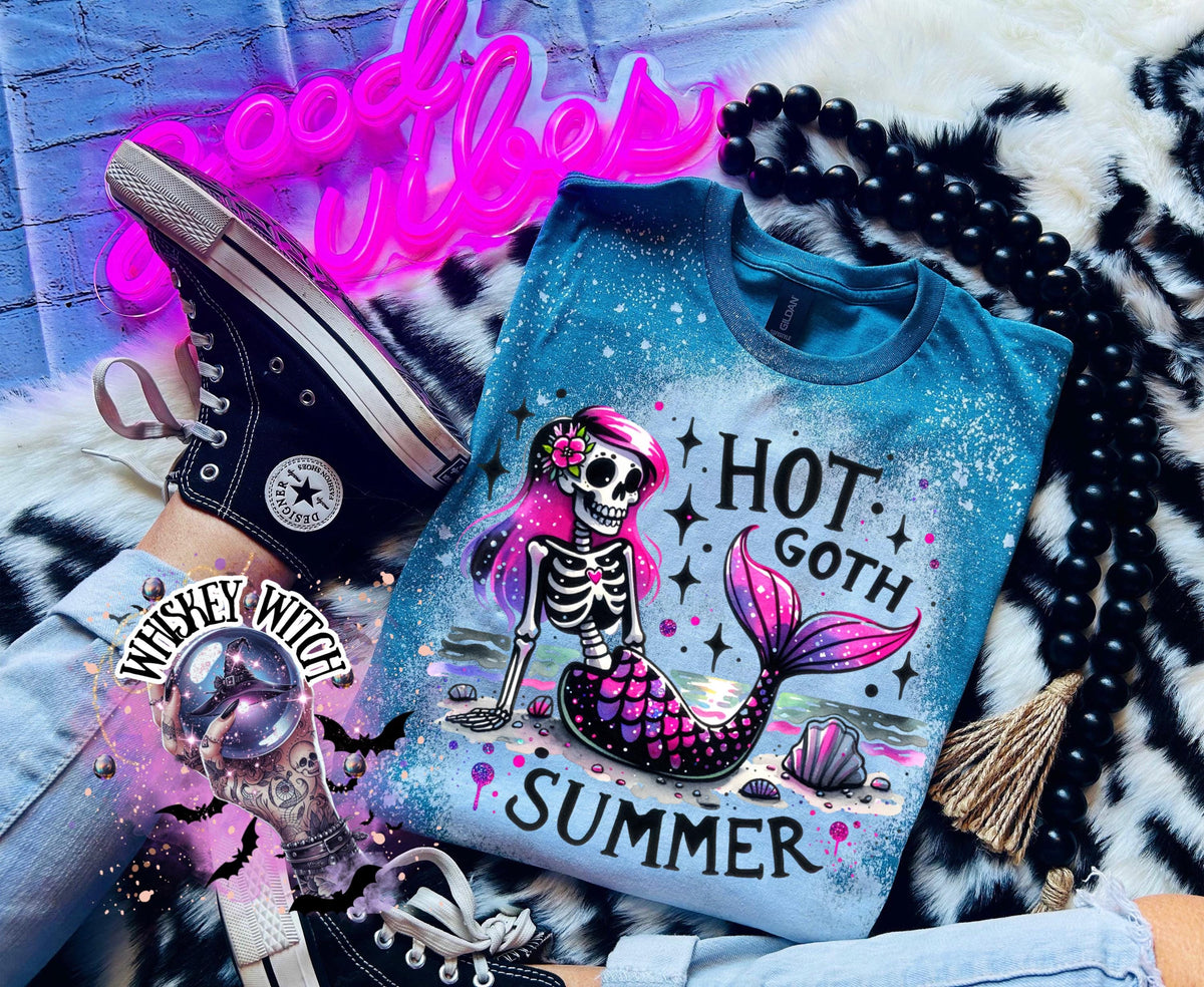 A teal colored  t-shirt with a bleach-splattered design featuring a pink and purple skeleton mermaid with the text "Hot Goth Summer".