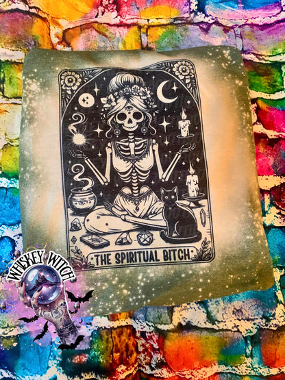 A bleached military green t-shirt with a black and white illustration of a skeleton woman with a cat and the text "The Spiritual Bitch".