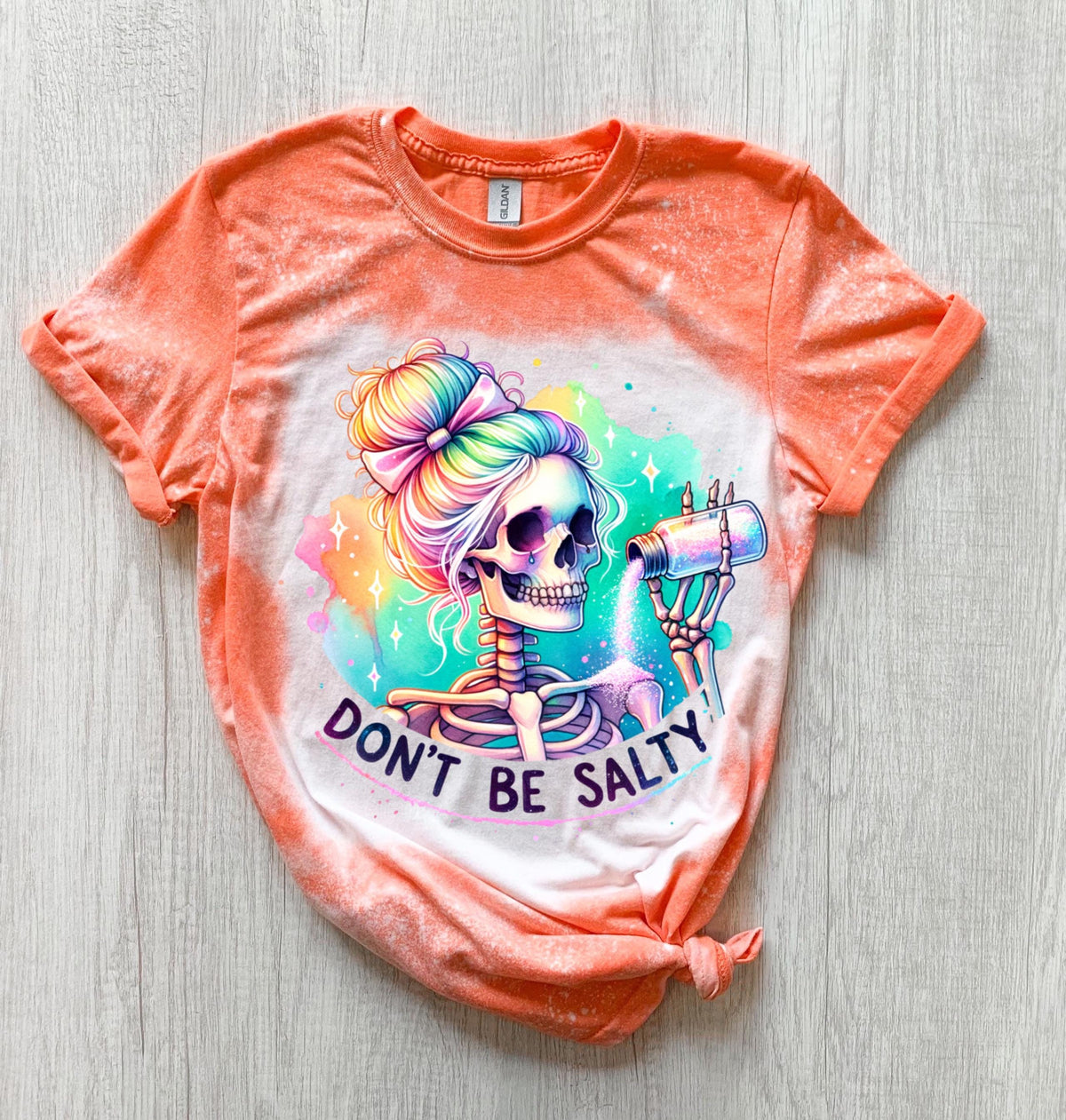 A Orange  t-shirt with a bleached effect featuring a skeleton with rainbow hair pouring salt from a shaker. The text "Don't Be Salty" is printed on the shirt