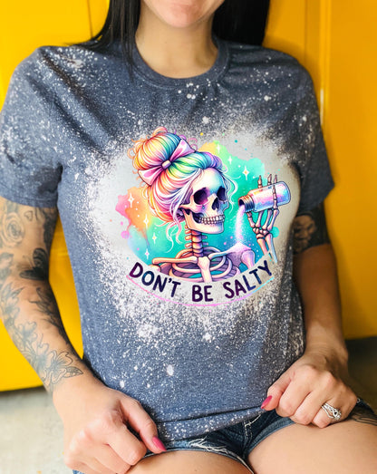 A navy blue  t-shirt with a bleached effect featuring a skeleton with rainbow hair pouring salt from a shaker. The text "Don't Be Salty" is printed on the shirt
