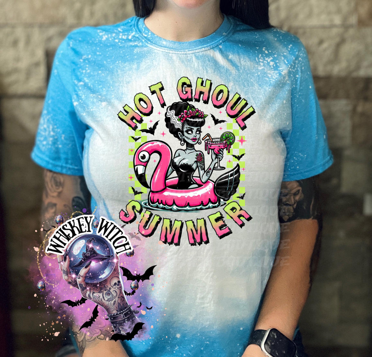 A sapphire blue  and bleach splattered effect  t-shirt with a graphic of a zombie woman in a flamingo pool float, holding a cocktail. The text "Hot Ghoul Summer" is printed on the shirt.