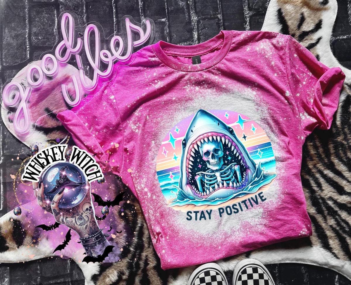 A pink t-shirt with a bleached design of a skeleton inside a shark's mouth. The shark is surrounded by a rainbow and stars. The text "Stay Positive" is below the shark.