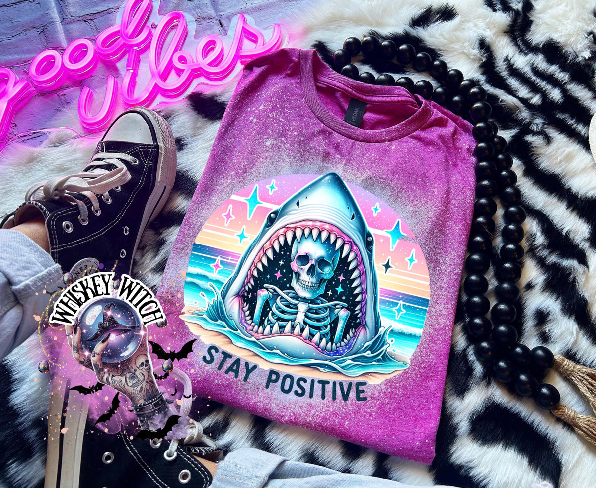 A Berry pink t-shirt with a bleached design of a skeleton inside a shark's mouth. The shark is surrounded by a rainbow and stars. The text "Stay Positive" is below the shark.