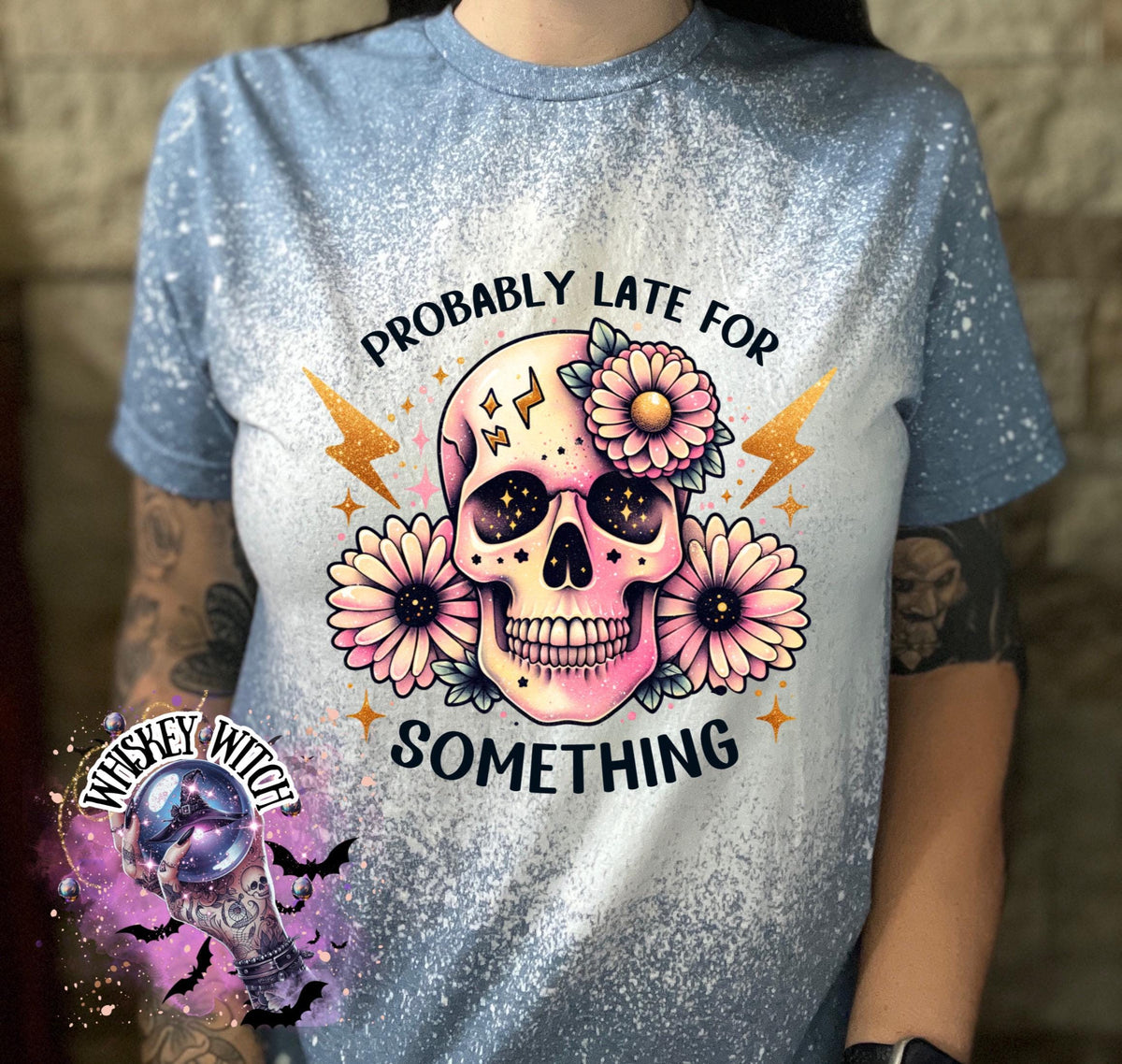 A indigo blue  t-shirt with a bleached effect featuring a skull with flowers and the text "Probably Late For Something" in gold glitter.