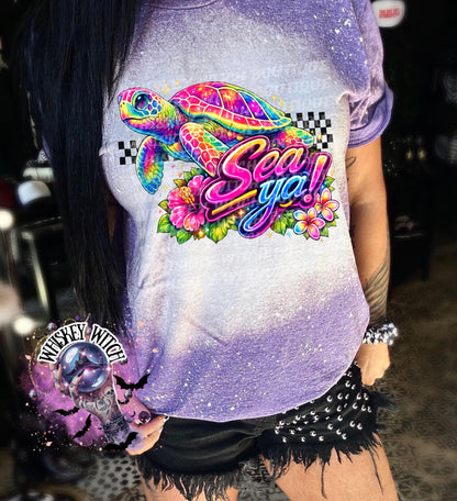 A purple t-shirt with bleach white effect a colorful graphic of a sea turtle, flowers, and the words "Sea Ya!". The shirt has a bleached, distressed look. The design features vibrant colors and a playful font.