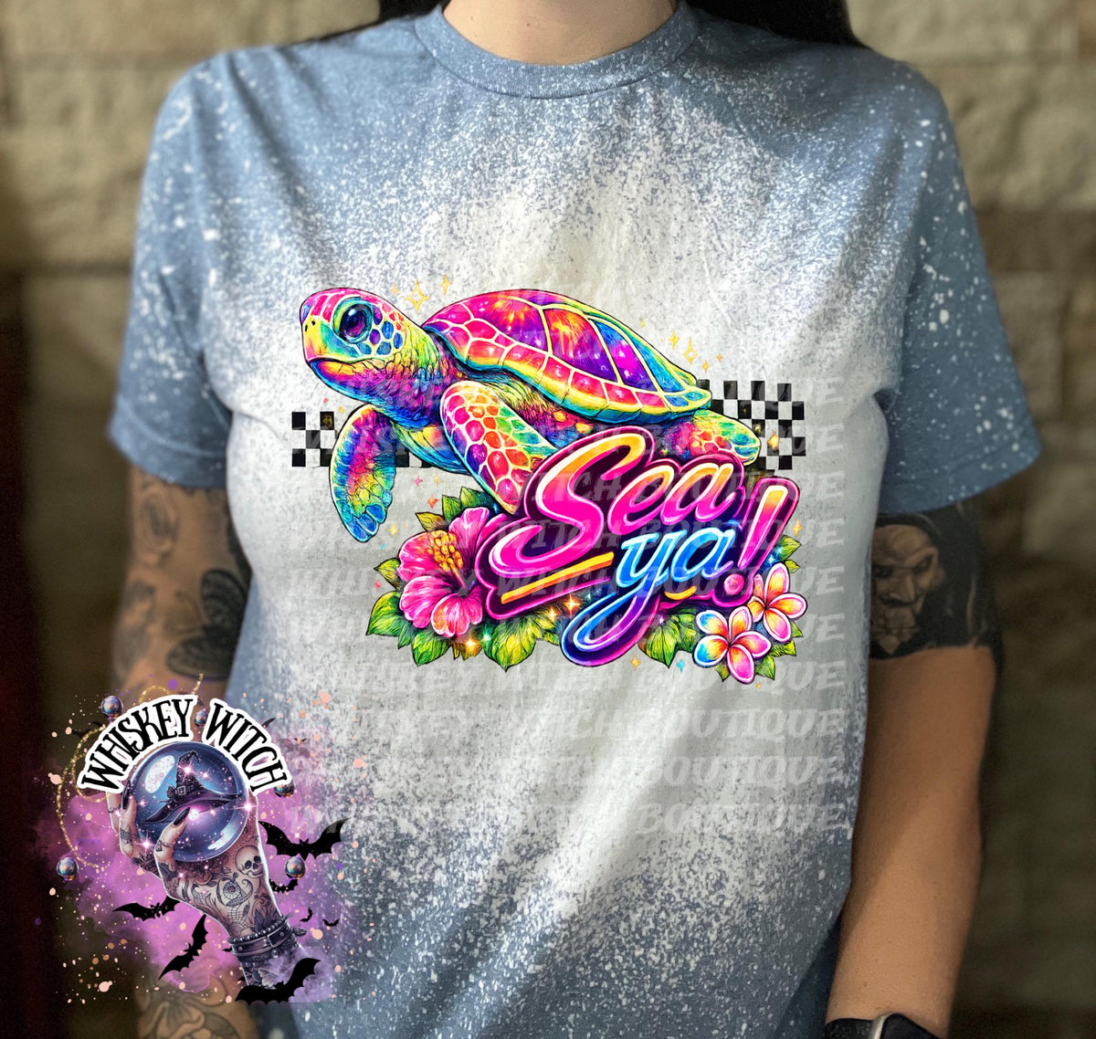 A indigo  blue t-shirt with bleach white effect a colorful graphic of a sea turtle, flowers, and the words "Sea Ya!". The shirt has a bleached, distressed look. The design features vibrant colors and a playful font.