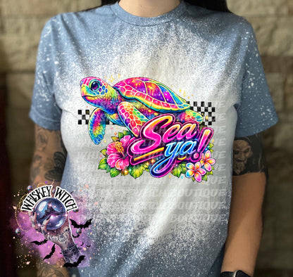 A indigo  blue t-shirt with bleach white effect a colorful graphic of a sea turtle, flowers, and the words "Sea Ya!". The shirt has a bleached, distressed look. The design features vibrant colors and a playful font.