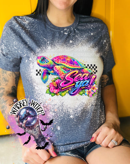 A navy  blue t-shirt with bleach white effect a colorful graphic of a sea turtle, flowers, and the words "Sea Ya!". The shirt has a bleached, distressed look. The design features vibrant colors and a playful font.