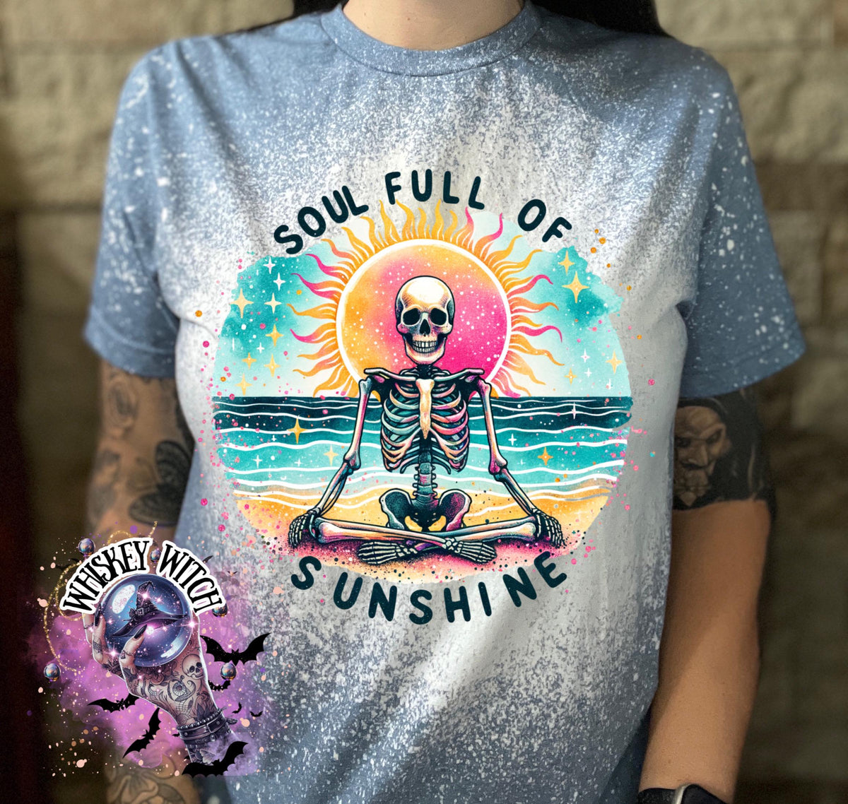 Light blue t-shirt with a bleached effect, featuring a colorful graphic of a skeleton meditating on a beach with a sun and the text "SOUL FULL OF SUNSHINE". The design includes turquoise, pink, yellow, and black elements.
