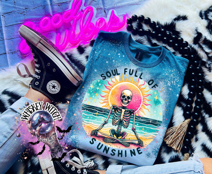 A Galapagos blue and bleach white splatter  t-shirt featuring a skeleton meditating on a beach with a sun in the background. The text on the shirt reads "SOUL FULL OF SUNSHINE".
