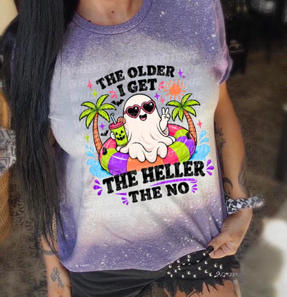A purple and white t-shirt with the text "The Older I Get The Heller The No". The shirt features a cartoon ghost wearing sunglasses and floating on a colorful inner tube, with palm trees and a drink.