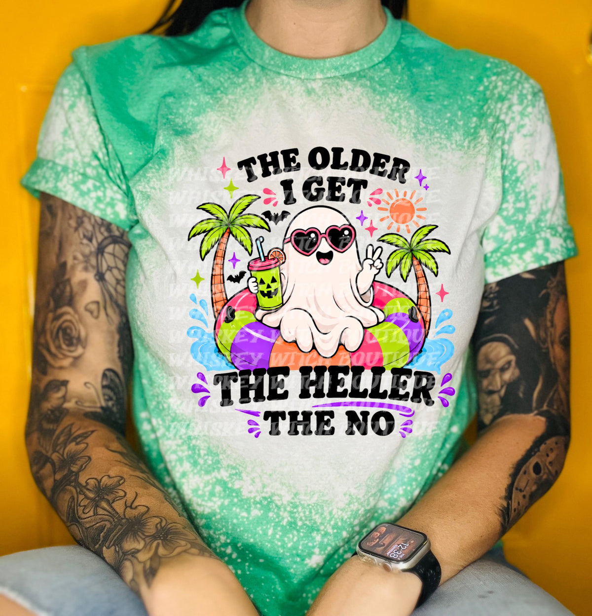A white t-shirt with a green The shirt features a cartoon ghost wearing heart-shaped sunglasses and a peace sign, floating on a colorful inflatable ring. Text on the shirt reads "THE OLDER I GET THE HELLER THE NO".