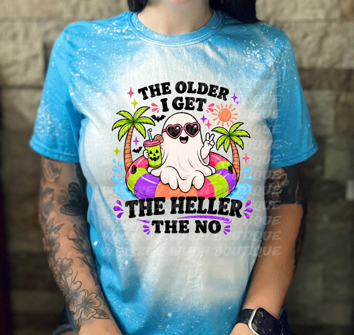 A blue and white tie-dye t-shirt featuring a cartoon ghost wearing sunglasses and floating on a colorful inner tube. The text on the shirt reads "THE OLDER I GET THE HELLER THE NO".