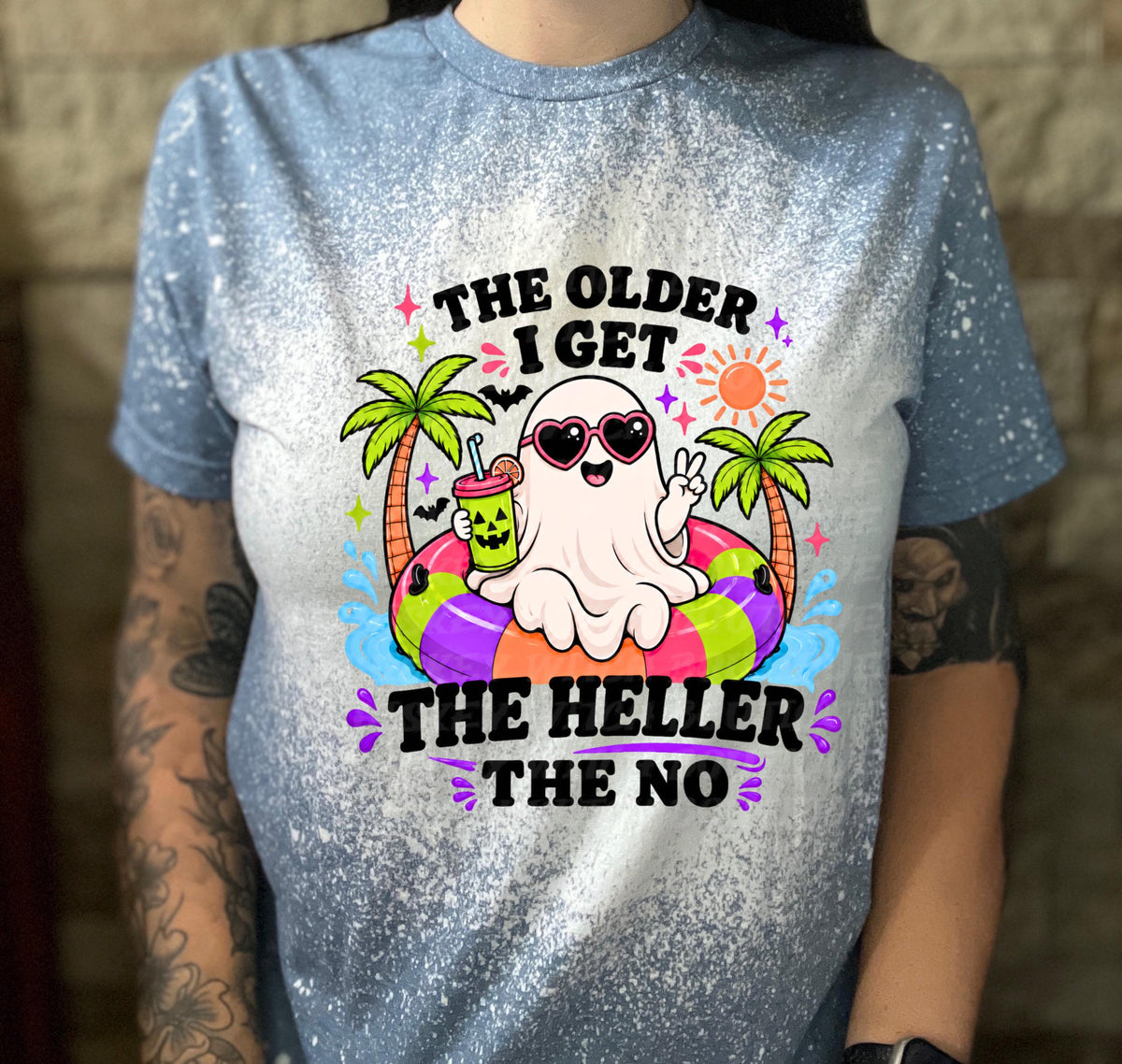 Light blue t-shirt with a distressed design. The shirt features a cartoon ghost wearing heart-shaped sunglasses and holding a drink, floating on a colorful inner tube. Text reads "THE OLDER I GET THE HELLER THE NO".
