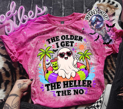 A pink t-shirt with a graphic of a ghost wearing sunglasses and floating on a colorful inflatable ring. The text on the shirt reads "The Older I Get The Heller The No". The shirt has a bleached, distressed look.