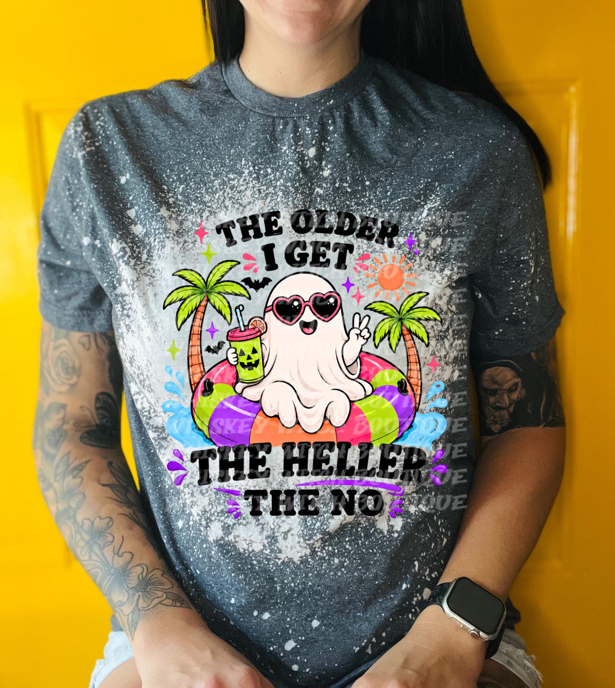 A gray t-shirt with a bleached design featuring a cartoon ghost wearing sunglasses and a pool float. The text reads "THE OLDER I GET THE HELLER THE NO". The ghost is holding a drink.